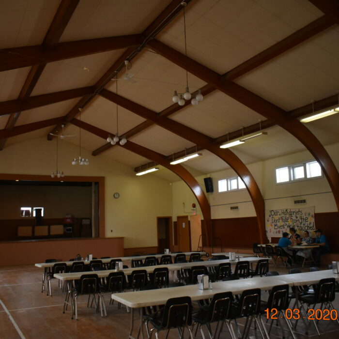 Colour photo of the interior of a hall with tables and chairs set up. The ceiling has large wooden beams and the walls are painted yellow. There are small windows which line the top half of the walls