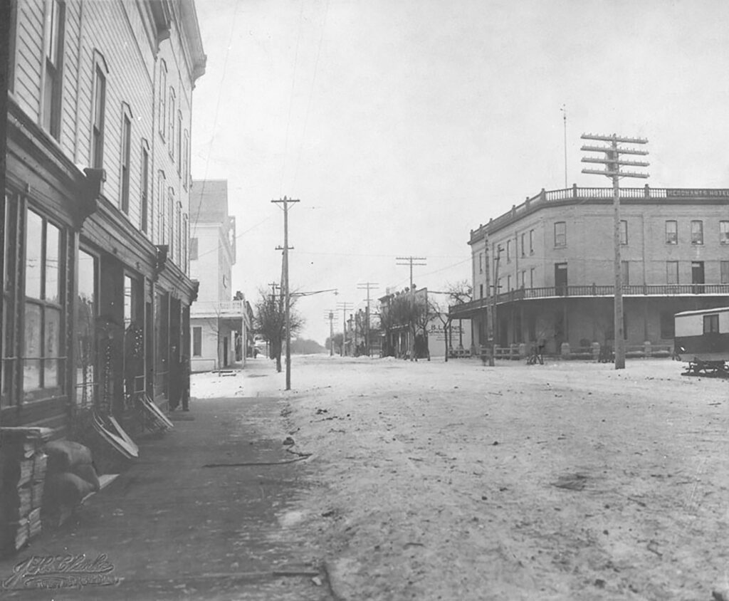 Black and white photo of the merchant's hotel from down the street. The street is unpaved and there are telephone poles along the sides of the road. On the left side of the street is a large multi-story building with big windows.