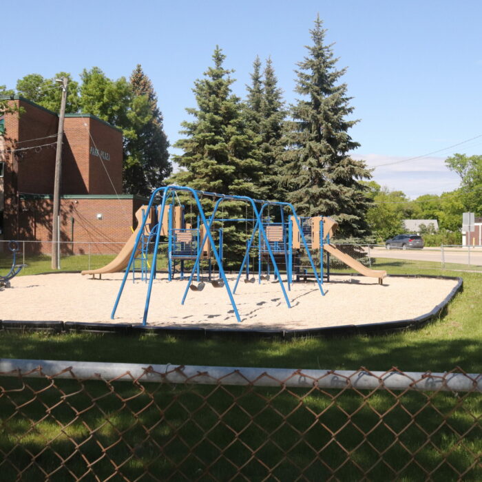 A colour image of a blue playground set on a gravel pad. There is grass around the playground with a chain link fence surrounding the space. In the background is a red brick building