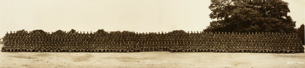 A long horizontal sepia-coloured image of a group of soldiers. There is a large tree on the right side of the photo. The men in the photo are so small and so many that its hard to see their faces