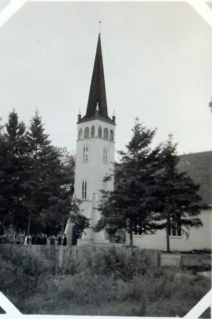 Black and white image of a white church with a tall steeple. There are trees on either side of the church