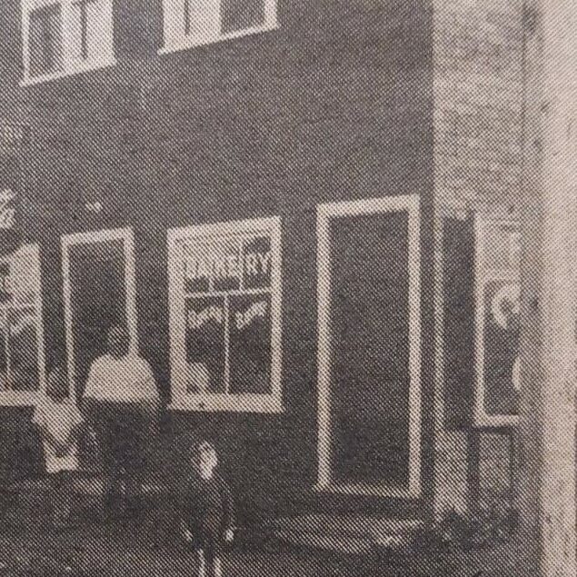 Original Selkirk Bakery on Manitoba Ave, Date Unknown, Selkirk Enterprise Centennial Edition Black and white photo of a two-story dark brick building. There are two windows and two doors at the front of the building and a man with two children