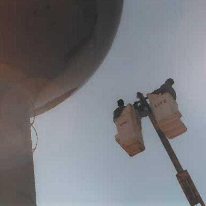 Colour photo of a crane with two buckets at the top of the crane. There is a man in either side of the crane. The crane is beside the base of the water tower
