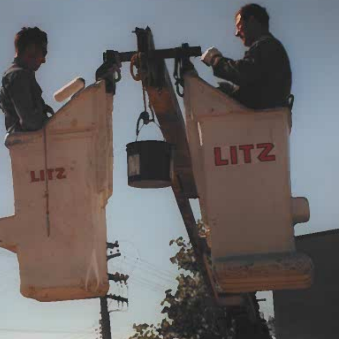 Colour photo of two men standing in t Litz crane. The crane has two buckets and have red letters which read 'litz' on the side