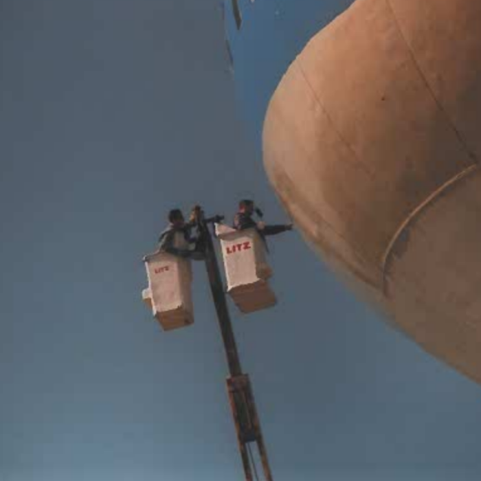 A colour image of painters in a crane raised up beside the water tower. One painter reaches out the side of the crane