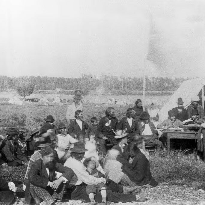 Black and white image of a large group of people gathered together. They are standing and seated on the ground with a tent in the background.