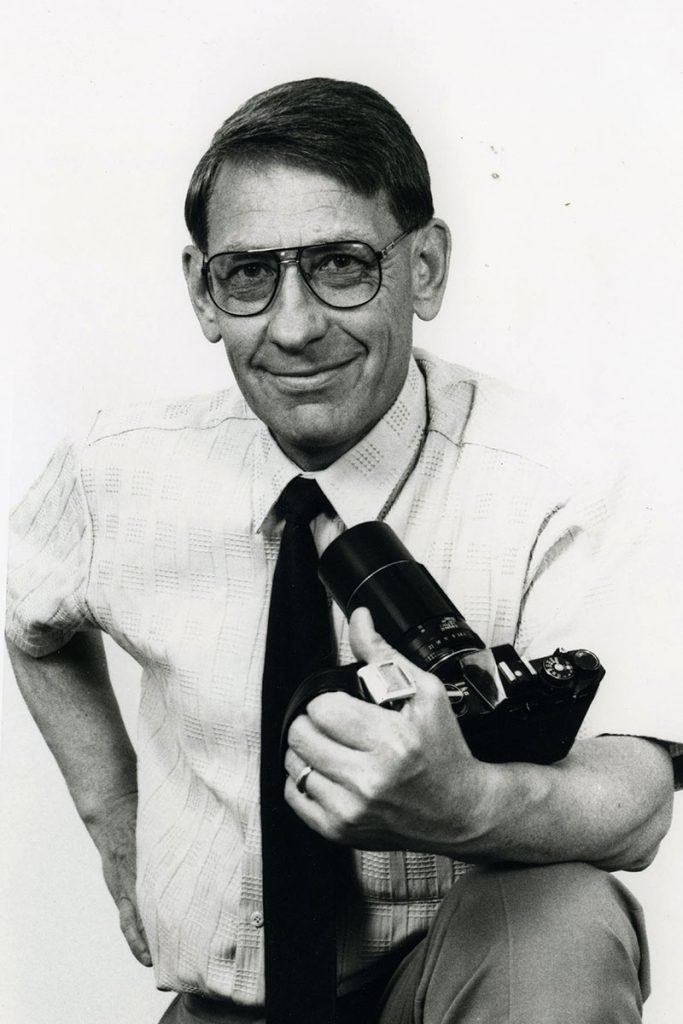 Black and white photo of man seated centrally in the frame. He is wearing a white button up shirt with a black tie, thick rimmed glasses, he is holding a camera and is smiling