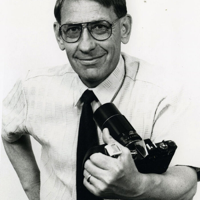 Podruchny Photography Instructor, 1973, Thomas Podruchny Black and white photo of man seated centrally in the frame. He is wearing a white button up shirt with a black tie, thick rimmed glasses, he is holding a camera and is smiling