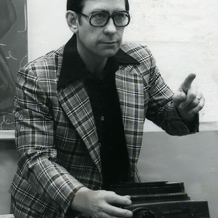 Podruchny Photography Instructor, 1977, Thomas Podruchny Black and white image of Tom wearing a plaid jacket, and dark thick rimmed glasses. He looks ahead and points