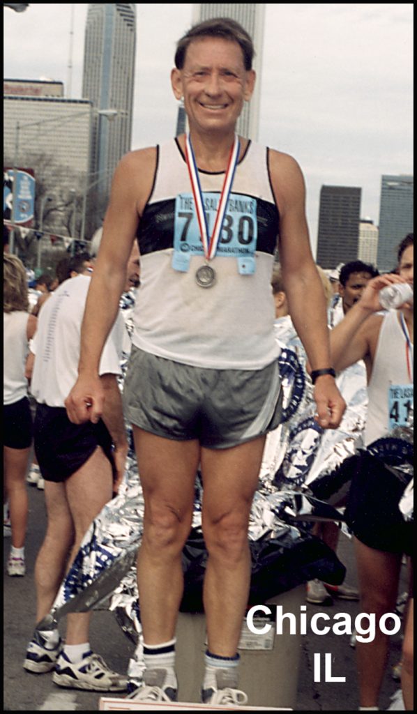 Man is standing with a silver medal around his neck. He is wearing a white and black shirt with grey shorts. He is smiling. The text 'Chicago IL' is printed in the bottom right corner