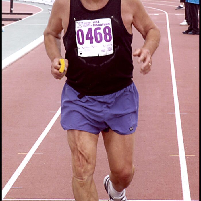 Podruchny at Manitoba, 2004, Thomas Podruchny Tom is running towards the camera. He is wearing a black shirt and purple shorts. The number 0468 is pinned to his shirt. He is smiling. Text 'Manitoba' is printed across the bottom right side