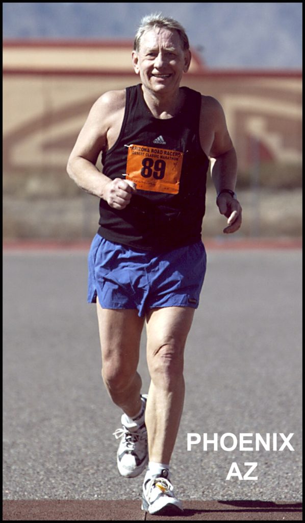 Man is positioned centrally in the frame running towards the camera. He is wearing a black shirt and blue shorts. The number 89 is pinned to his shirt. He is smiling