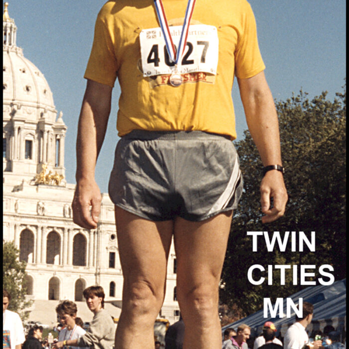 Podruchny at Twin Cities, 1993, Thomas Podruchny Tom stands centrally in the frame wearing a yellow shirt and grey shorts. e has the number 4027 pinned to his shirt. In the background is a white building and people
