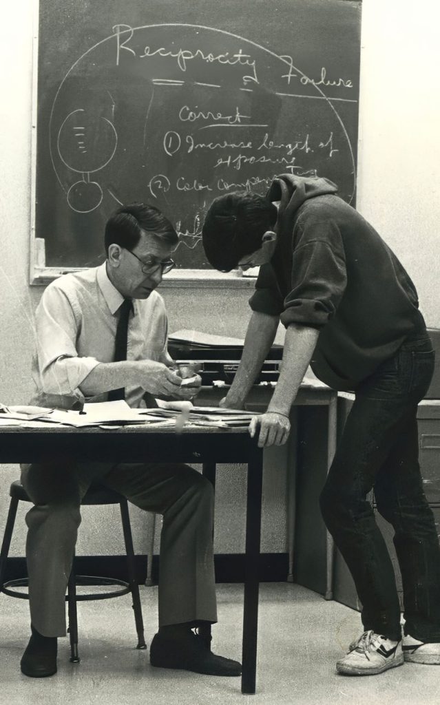 Black and white photo of man wearing white bbutton up shirt and black tie seated at a desk. A student stands opposite him and leans over the table