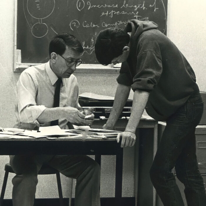 Podruchny Photography at the Comp, 1974, Thomas Podruchny Black and white photo of man wearing white button up shirt and black tie seated at a desk. A student stands opposite him and leans over the table