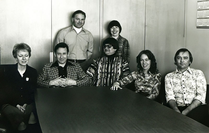 Black and white image of a group of adults standing and sitting against a wall