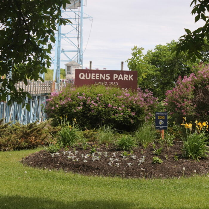 Queen's Park, 2020, Selkirk Museum Collection A colour image of a grass space with designated areas for flowers and ornamental grasses. In the background are trees and a wooden sign which reads 'Queens Park'. In the far distance is the Selkirk Lift Bridge