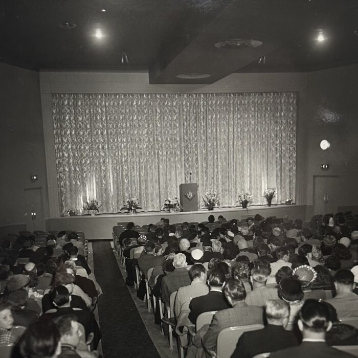 A black and white photo of the interior of the Garry Theatre filled with people. the curtain is drawn and the lights are dim