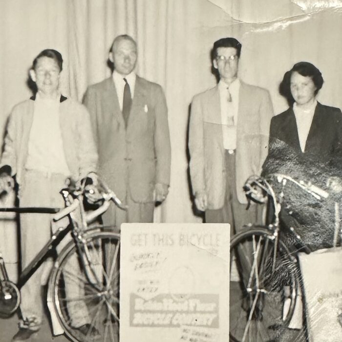 A black and white photo of a group of people standing on the stage of the Garry Theatre. There is a woman on the right. The group stands in front of a few signs