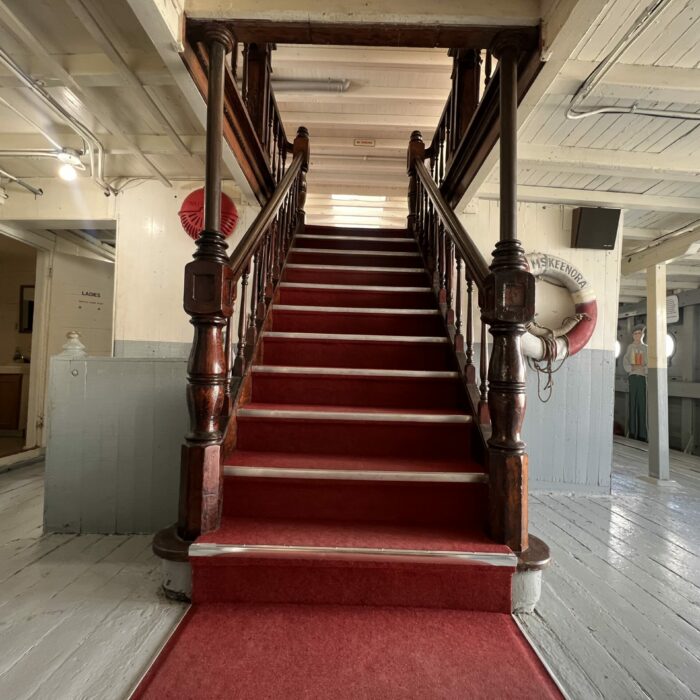 S. S. Keenora Staircase, 2026, Selkirk Museum Collection Colour photo of the main staircase in the Keenora. The staircase is lined with red carpet and the railing is wood.