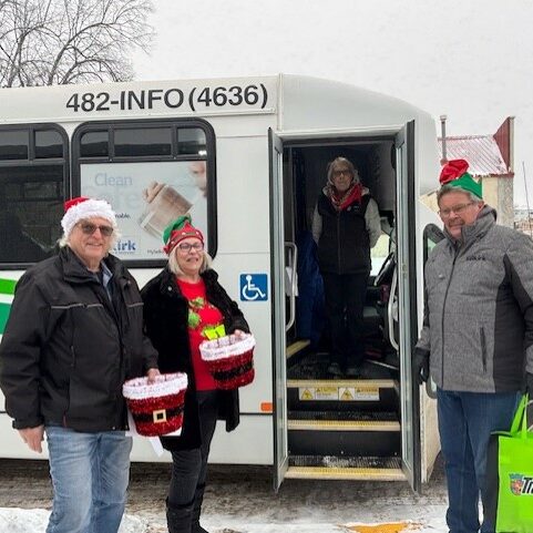 Colour photo of a bus parked in the snow. The door is open and the bus driver is visible. standing in front of the door are three people wearing Santa hats and holding Christmas decor