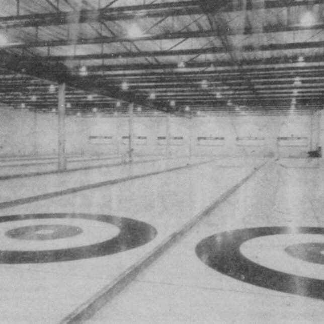 Black and white image of a curling rink. The room is large with multiple lanes, a high ceiling, and lots of bright lights
