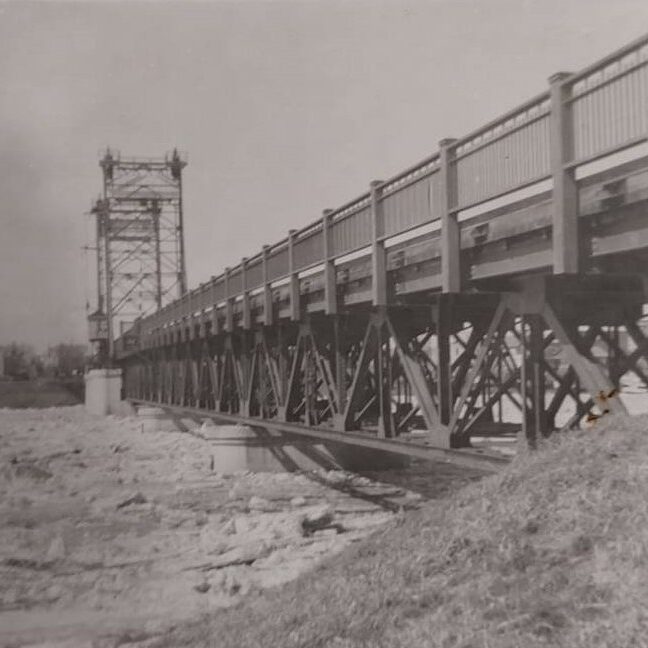 A black and white image of a bridge spanning a frozen river. The photo is taken from the bank on one side of the bridge looking down the length of it