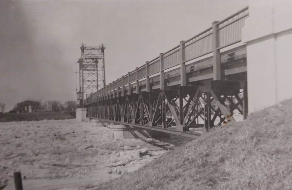 A black and white image of a bridge spanning a frozen river. The photo is taken from the bank on one side of the bridge looking down the length of it