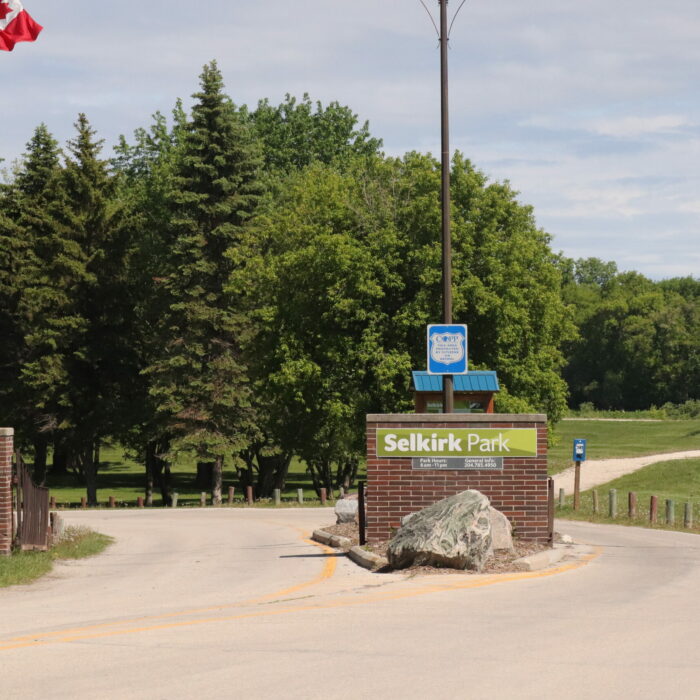 Selkirk Park, 2020, Selkirk Museum Collection A colour image of the entrance to Selkirk Park. There are trees in the background, a gravel walking path off to the right hand side. In the foreground is the paved road leading into the park, a brick sign which reads 'Selkirk Park' and a flag pole with the Canadian flag flying