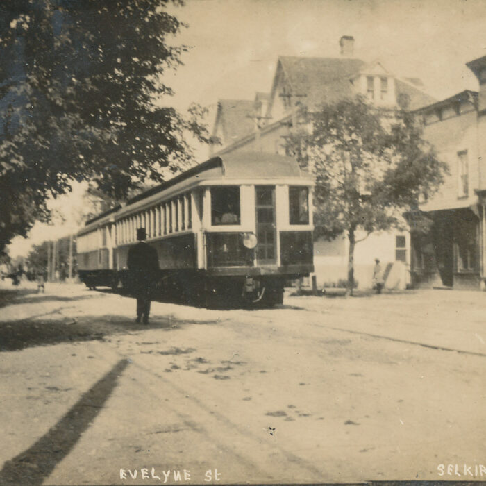 Black and white image of a street with a train car. A man stands to the left of the streetcar and on the right side of the street car are buildings and a small tree. There is a tree on the left of the image as well