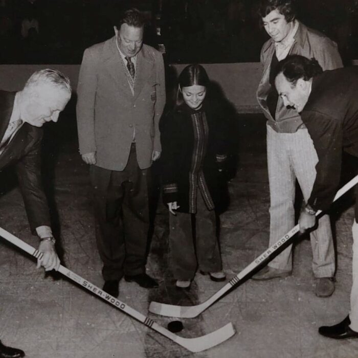 Woman drops the puck at the center of the image. On either side of her stands two men. In the foreground two men smile holding hockey sticks ready to play