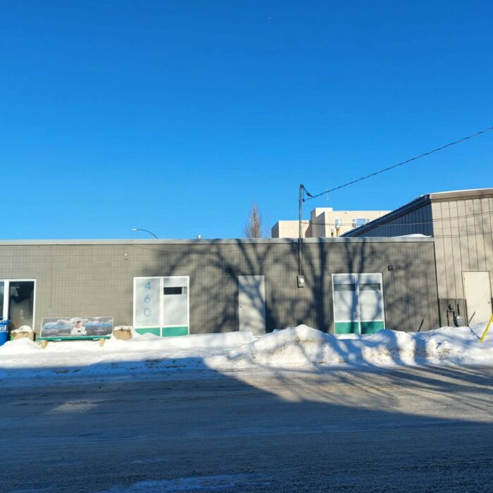 Colour image of a long rectangular grey building at the corner of an intersection. On the right side of the building is a large garage