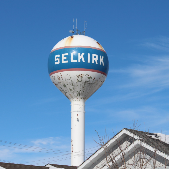 A colour photo of the selkirk water tower. The tower is behind the peaks of a few houses. The white paint on the tower is peeling