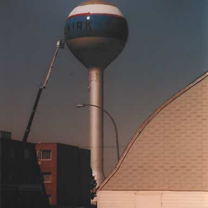 A colour image of the water tower at dusk. In the foreground is the corner of the selkirk arena. On the left side of the frame is the crane with the painters