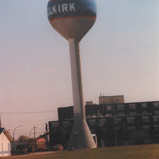 Colour photo of the Selkirk Water tower from across the field. In the background is a multi-story building.