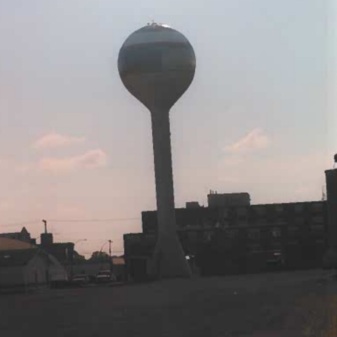 A colour image of the water tower from across the field. In the background are buildings. The sun is setting and the sky is pink and blue.