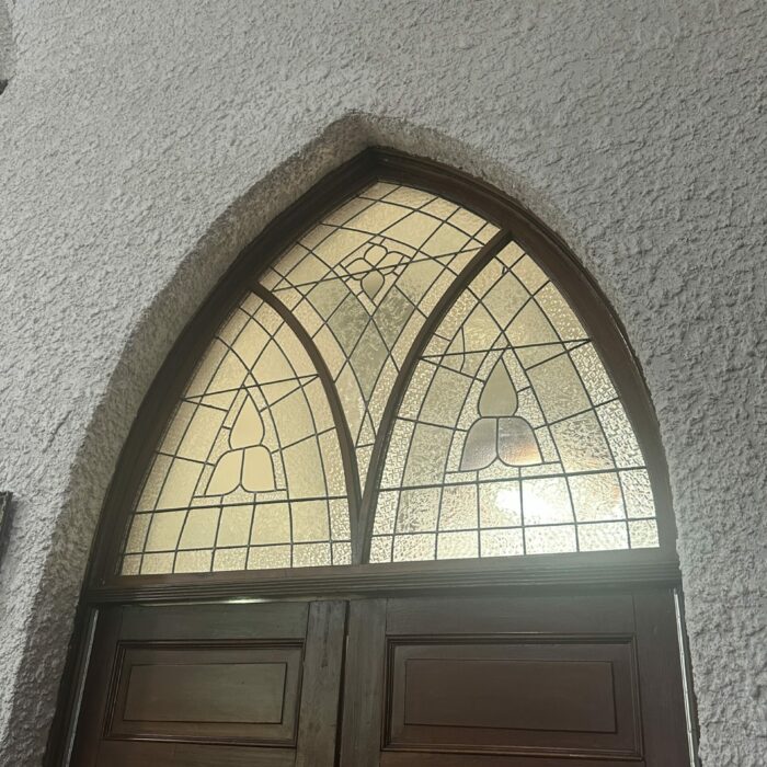 A colour photo of the top of a wooden door. There is a stained glass panel and stucco walls
