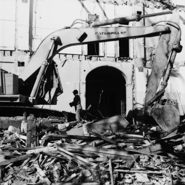 The Demolition of the Selkirk Mental Hospital 1, Date Unknown, Madness Canada Black and white photo of a large excavator tearing down a building. There is rubble on the ground surrounding the excavator and an archway with a man standing beside it in the background