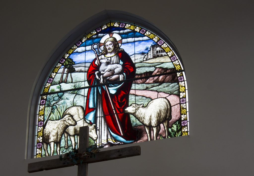 Stained glass window showing image of Christ gathering his flock. He is wearing a red robe with a blue robe underneath. He is holding a lamb with three sheep at his feet