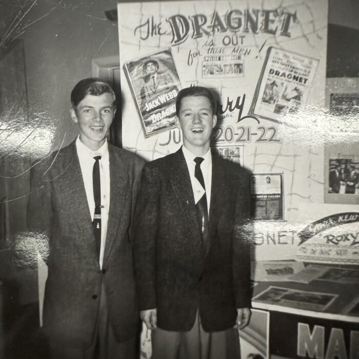 A black and white photo of two young men standing together in front of a sign which reads 'dragnet'. The two men wear suits and ties