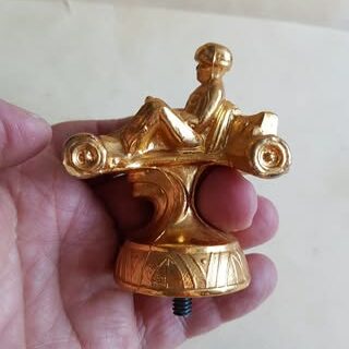 Colour image of the top part of a gold trophy. The trophy topper is in the shape of a child driving a small derby car