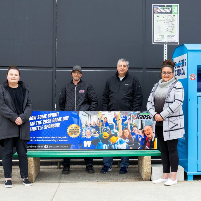Colour image of four people standing at a bus stop. They stand around a bench