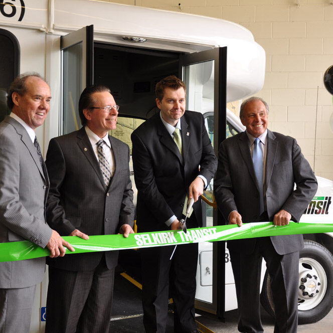 This is a photo of CAO Duane Nicole cutting a green ribbon with "Selkirk Transit" spelled across the front. Duane Nicole is alongside local NDP leader Greg Dewar, and Mayor Larry Johannson. They are all dressed nicely in suits, in front of a City of Selkirk transit bus.