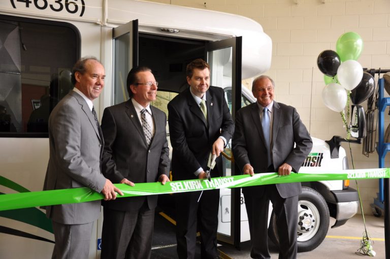 This is a photo of CAO Duane Nicole cutting a green ribbon with "Selkirk Transit" spelled across the front. Duane Nicole is alongside local NDP leader Greg Dewar, and Mayor Larry Johannson. They are all dressed nicely in suits, in front of a City of Selkirk transit bus.