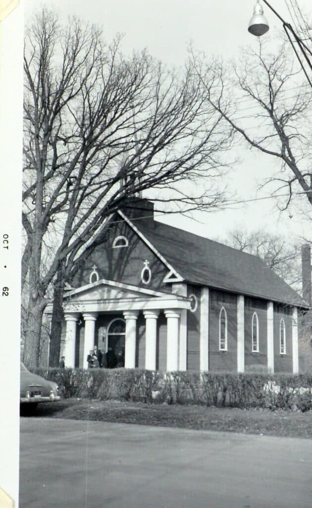 Photo of Selkirk Notre Dame Church. Photo shows off the front of the building that has an A-frame roof and pillars on either side of the front door. The sides if the church have pillars and windows between each window.