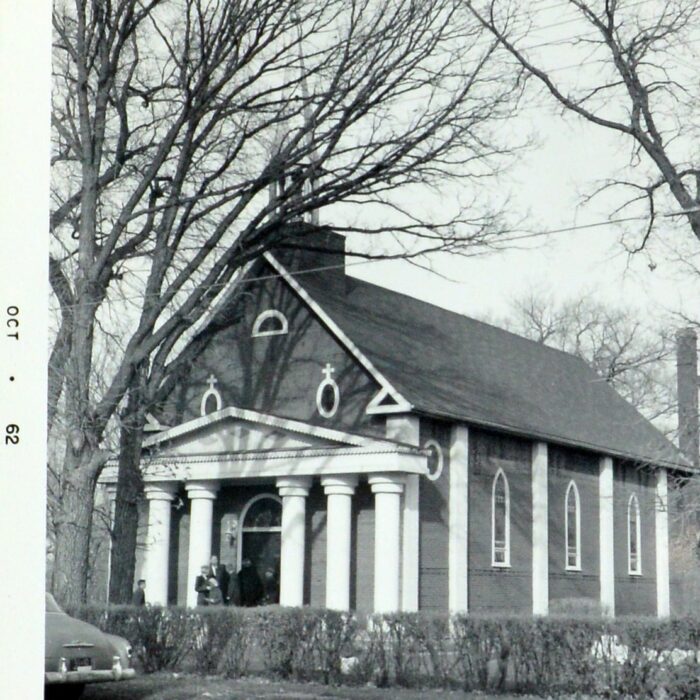 Photo of Selkirk Notre Dame Church. Photo shows off the front of the building that has an A-frame roof and pillars on either side of the front door. The sides if the church have pillars and windows between each window.