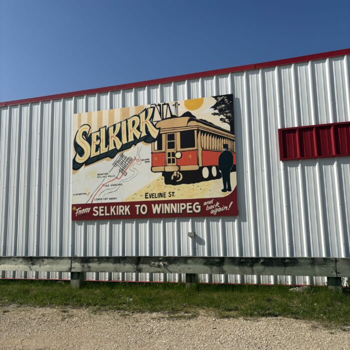Colour photo of a white tin sided building with red trim. There is a large sign which reads 'Selkirk' and a train car