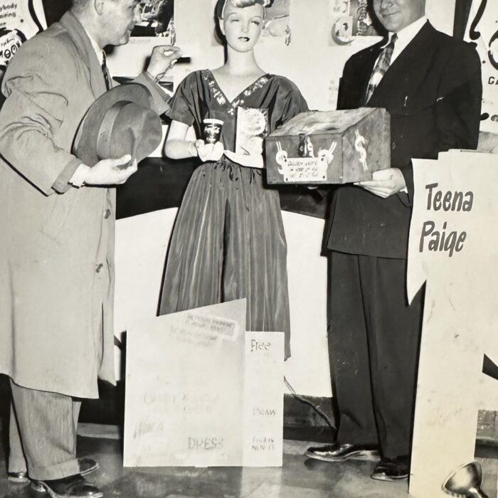 A black and white photo of a group of people. The woman at the center of the photo wears a long dress. The man on the left holds a top hat and wears a long trench coat. The man on the right wears a suit and tie and is holding a box. There is a post on the right side of the photo with the words 'Teena Paige'