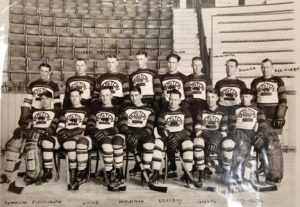 Black and white group photo of hockey players standing and kneeling on the ice surface. Bleachers in background