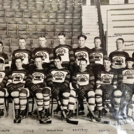 Black and white group photo of hockey players standing and kneeling on the ice surface. Bleachers in background